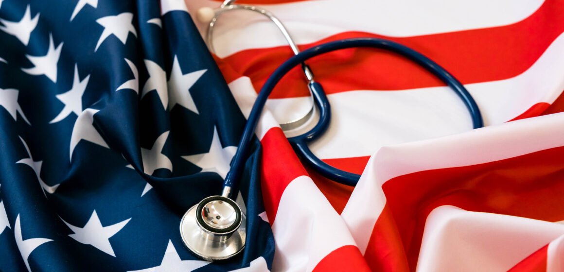 Close-up Photo Of Stethoscope On American Flag. medicine USA A stethoscope rests on a fabric styled like the American flag, with stars and stripes visible. The image is used on the article to show US Healthcare