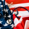 A stethoscope rests on a fabric styled like the American flag, with stars and stripes visible. The image is used on the article to show US Healthcare